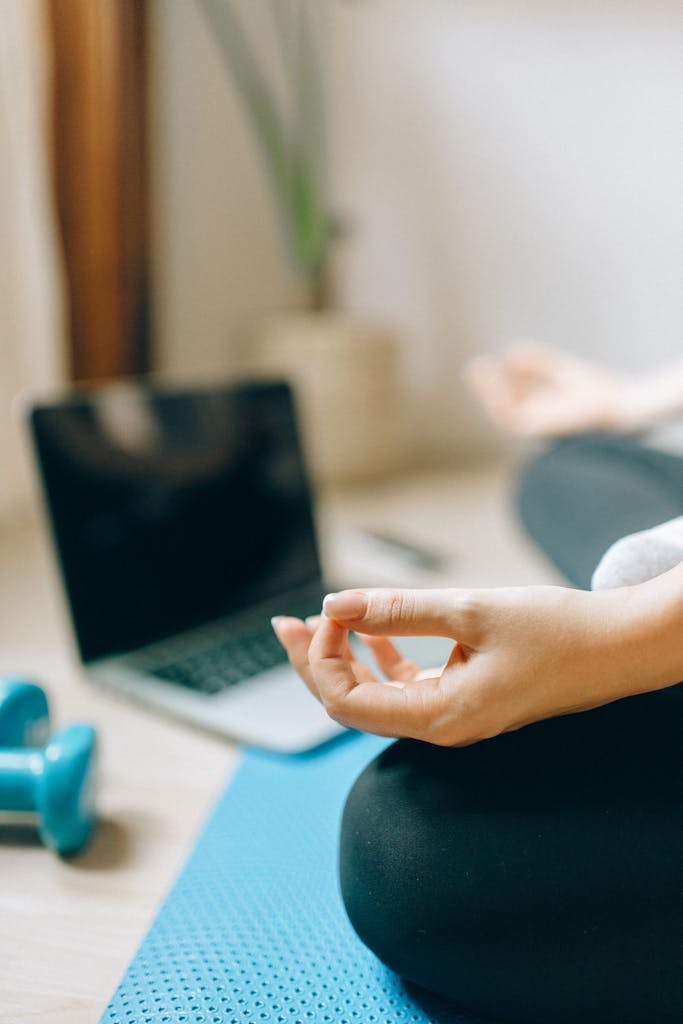 Adult woman meditating on yoga mat next to laptop and dumbbells indoors, promoting health and relaxation.